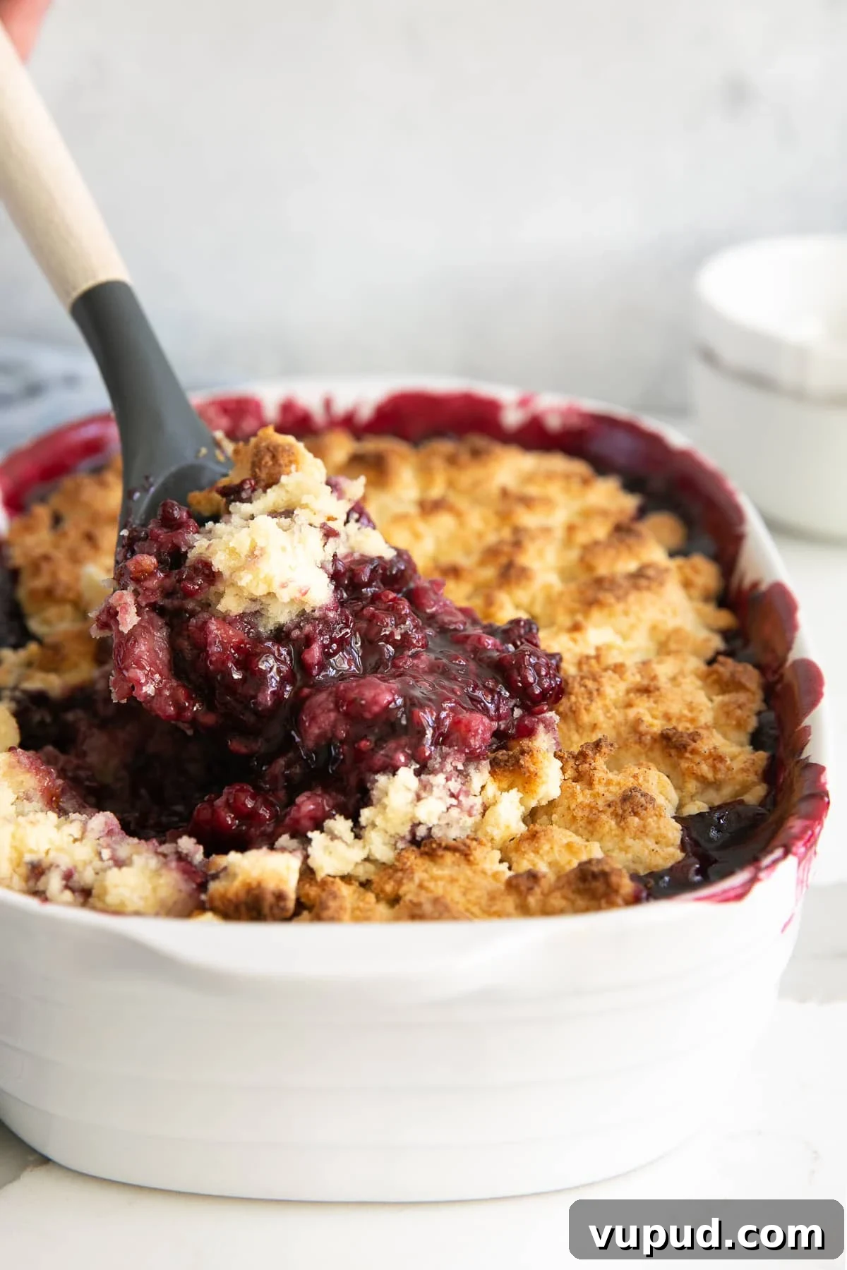 Classic Blackberry Cobbler Recipe: Sweet, Juicy, Homemade Dessert 2 Spatula scooping hot blackberry cobbler from a white baking dish.