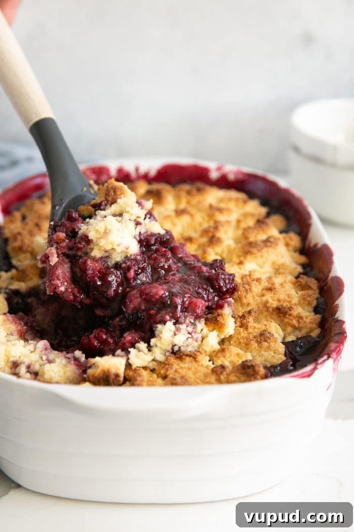 Spatula scooping hot blackberry cobbler from a white baking dish.
