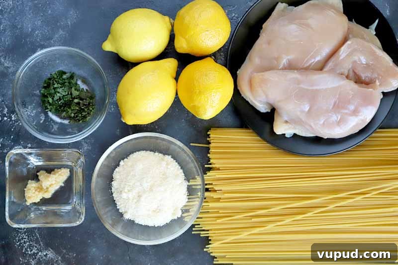 Overhead horizontal image of two round glass bowls of minced herbs and grated parmesan cheese, a small square dish of minced garlic, four lemons, a black plate of boneless, skinless chicken breasts, and a pile of uncooked spaghetti, on a mottled dark gray surface.
