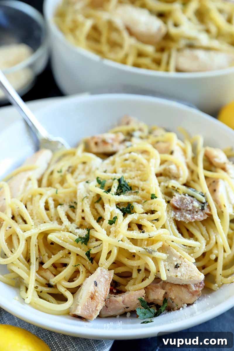 Vertical image of a white bowl of spaghetti with chicken and herbs, with a fork, with a white serving dish of more of the entree in the background beside a small glass dish of grated cheese, with two lemons, on a gray surface.