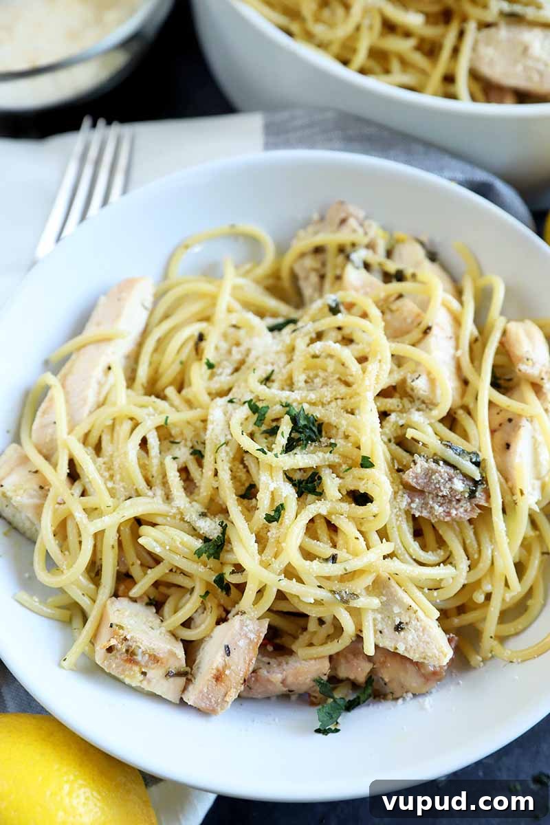 Vertical image of a white bowl of spaghetti with grilled poultry and herbs, on a gray background with a fork, a serving bowl filled with the dish, a small glass bowl of grated cheese, and a lemon, on a dark gray surface.