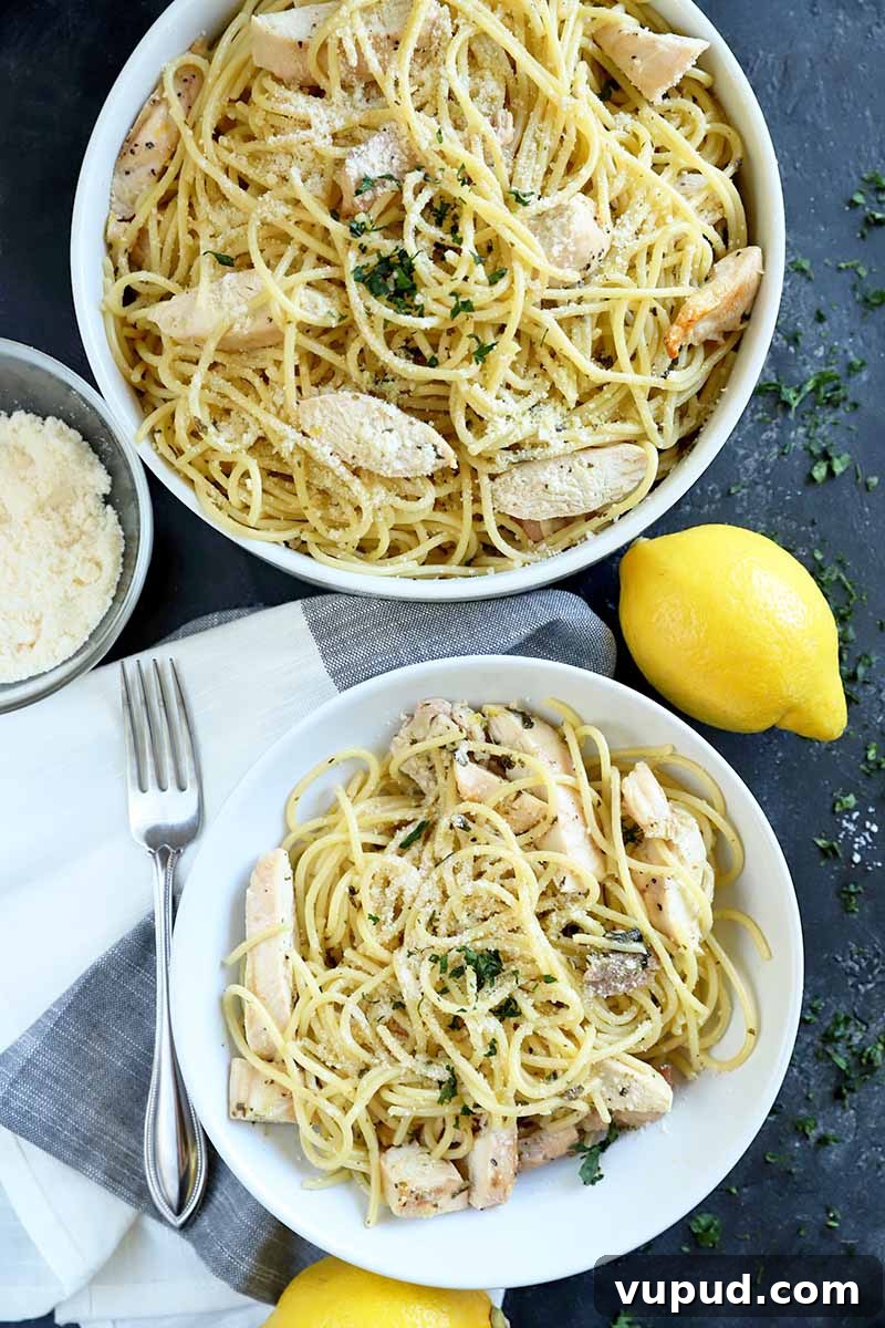 Overhead vertical image of a white ceramic bowl and a matching serving dish of spaghetti with sliced grilled chicken and herbs, on a gray surface topped partially with a white and gray cloth, with a small glass bowl of grated cheese, a fork, and two lemons.
