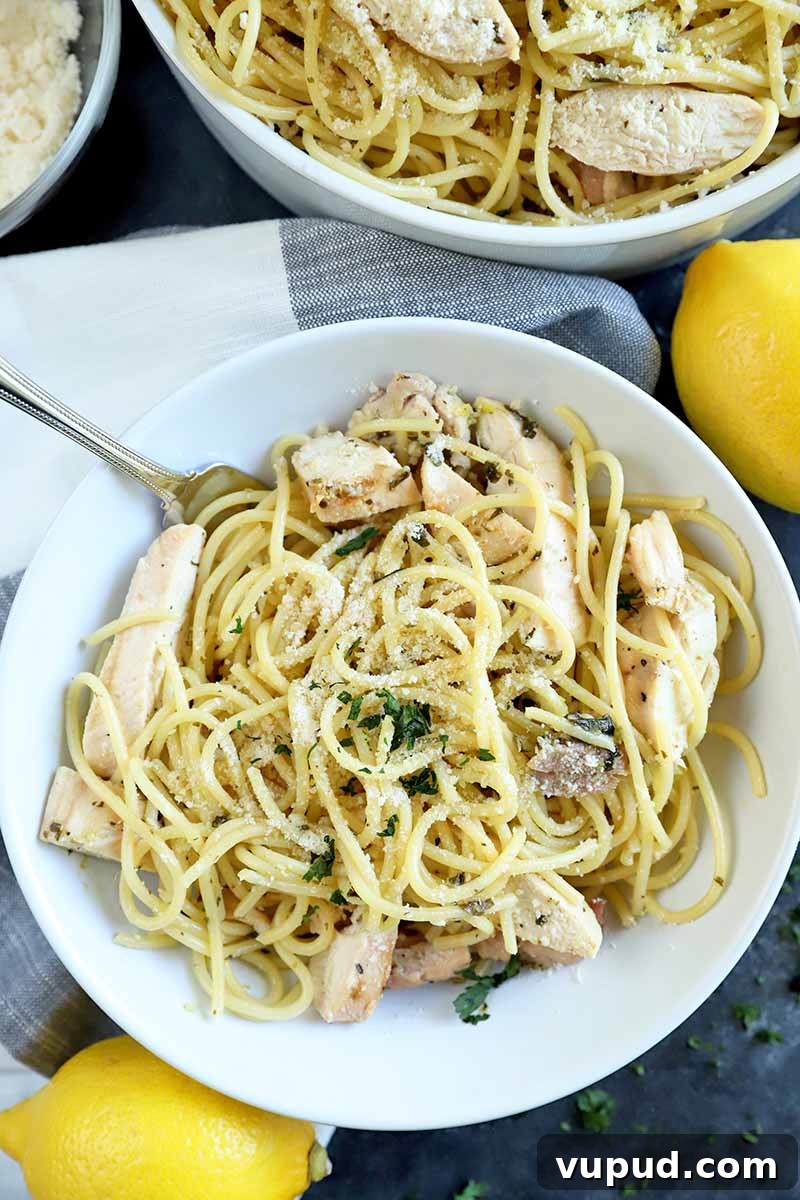 Vertical overhead image of a white bowl and larger white serving bowl of pasta with grilled poultry and herbs, with a fork, next to a small dish of grated cheese and two lemons, on a gray surface topped with a gray and white cloth.
