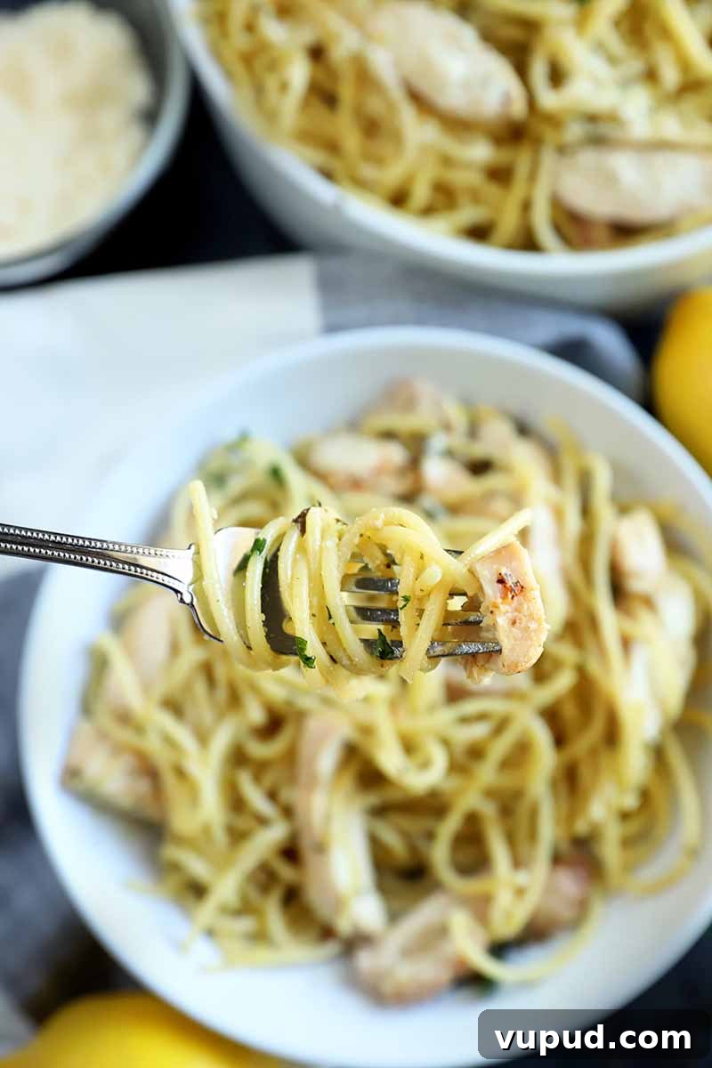 Vertical image of a fork holding a bite of spaghetti up to the camera, with more in a white bowl and a serving dish in the background, alongside a small glass dish of grated cheese and two lemons, on a white and gray cloth on top of a dark gray surface.