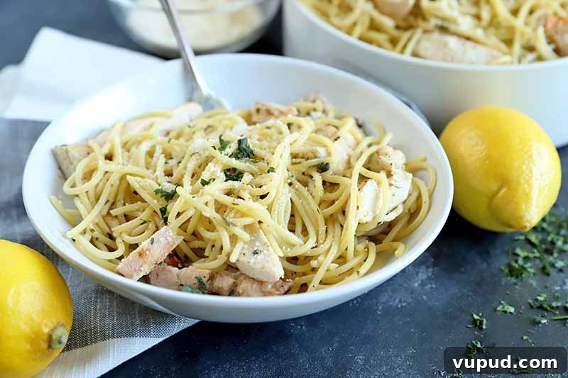 Horizontal image of a white bowl of spaghetti with sliced grilled poultry and chopped herbs on top, with a white ceramic serving bowl containing more of the dish in the background, next to a small glass dish of grated cheese, on a white and gray folded cloth with two whole lemons and scattered herbs, on a gray surface.