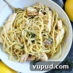 Overhead closely cropped horizontal image of a bowl of spaghetti with chicken, with two lemons on a white and gray cloth, on top of a dark gray surface.