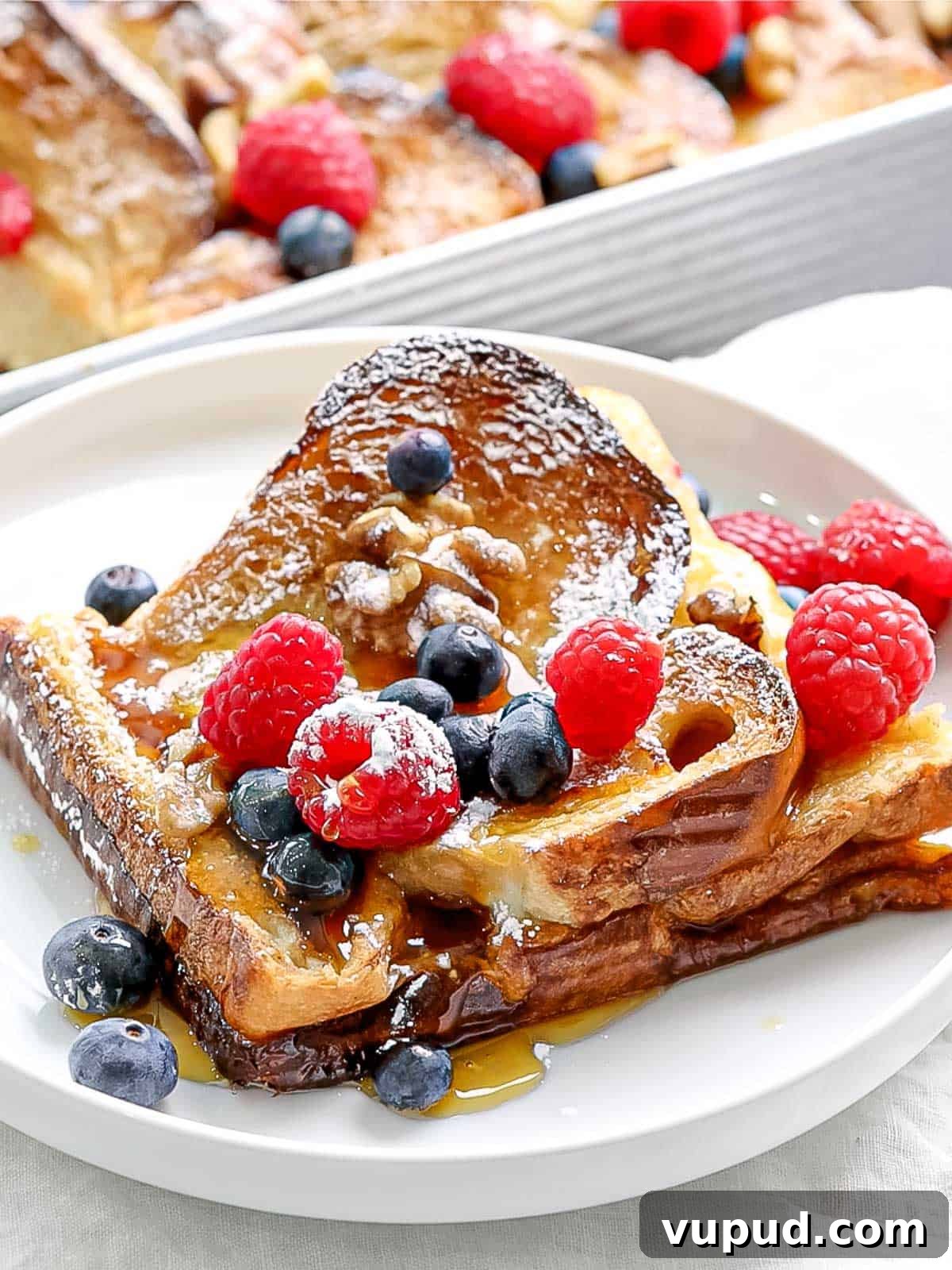 Make-Ahead Baked French Toast Casserole Recipe 8 Slice of baked french toast with blueberries, raspberries and powdered sugar on a white plate.