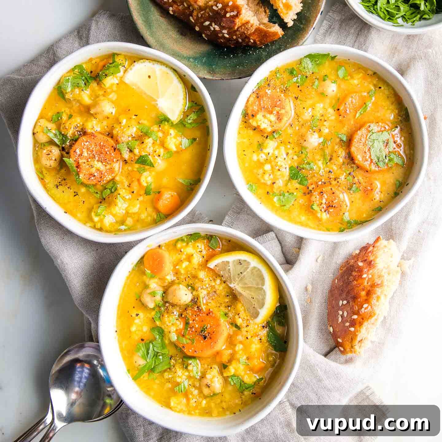 Lemony Vegan Chickpea and Rice Soup Recipe 10 Three bowls, filled with chickpea, rice, lemon and carrot soup, some crusts of bread lying around on a linen napkin with spoons on the side.