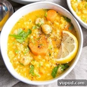 Lemony Vegan Chickpea and Rice Soup Recipe 11 White bowl filled with thick soup. Bright yellow broth with chickpeas, rice, carrots, lemon wedge and sprinkled with chopped parsley, salt and pepper.